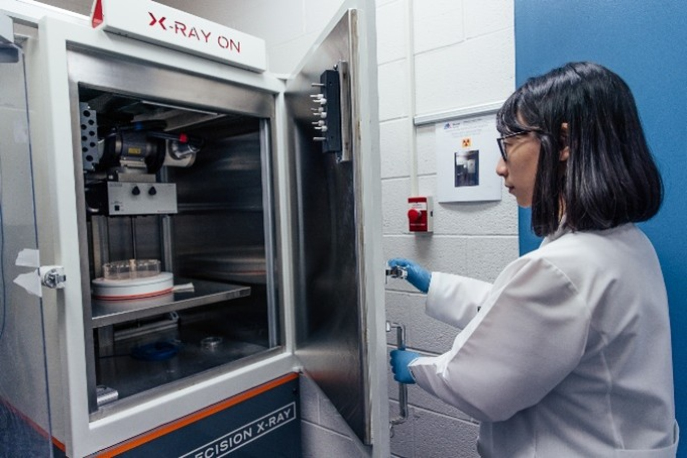 A woman opens the door to a machine that has the words X-RAY ON written above it -- but not lit up. It's shaped like small refrigerator and the door opens the same way. She is wearing a white lab coat and blue gloves.