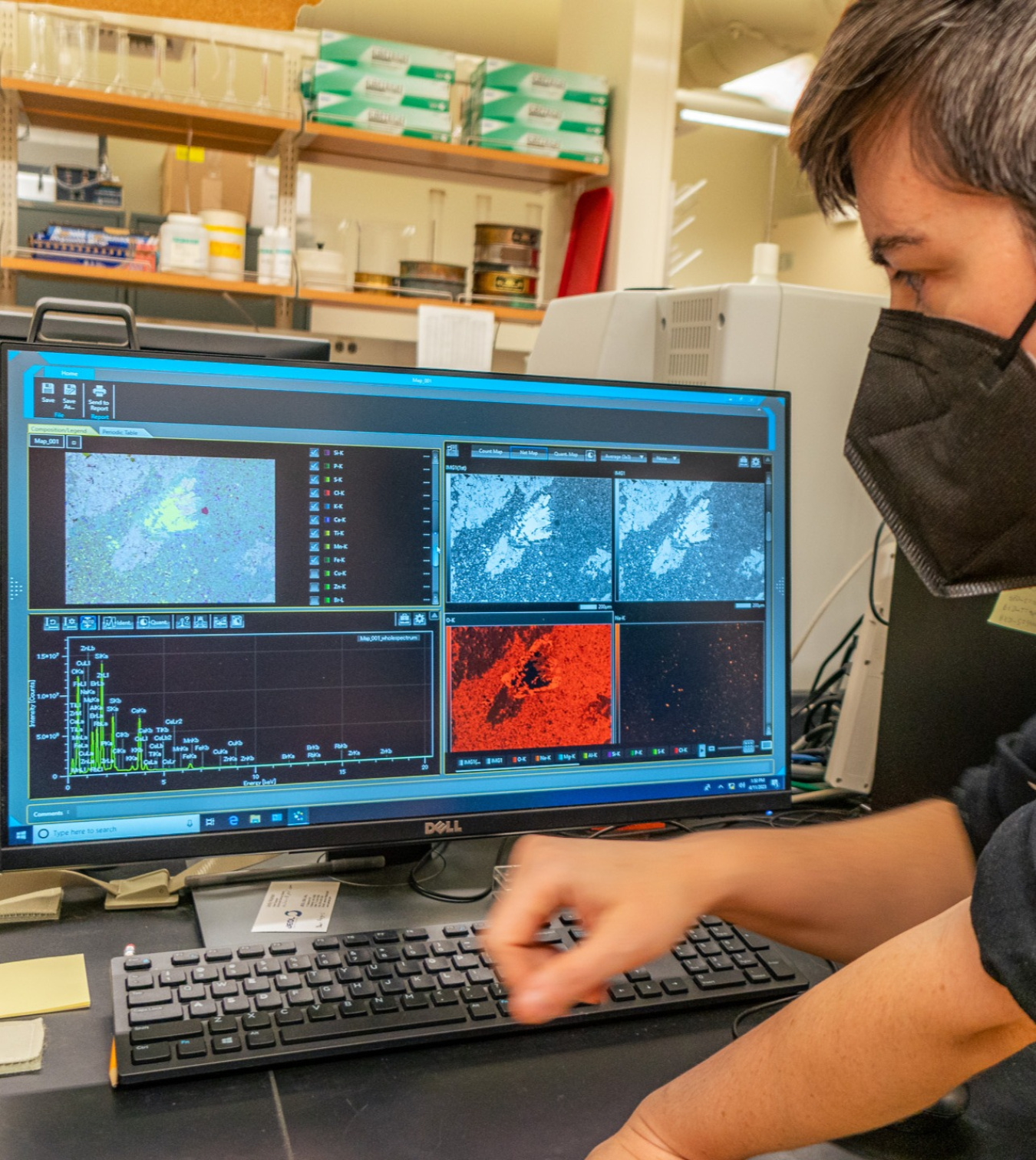 Computer screen displaying data from a geothermal project in a lab, while a scientist wearing a mask looks on