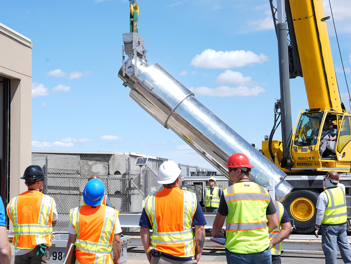 A crane holds a tall silver cylinder while 5 workers in reflective vests and helmets lookon.