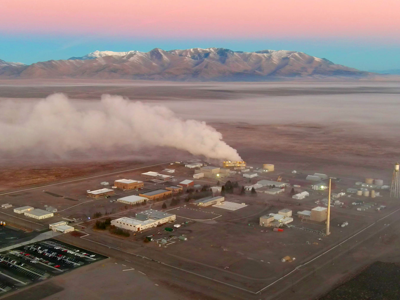Aerial view of Idaho National Lab with mountains and pink clouds in the background and a tall plume of steam rising from the Advanced Test Reactor