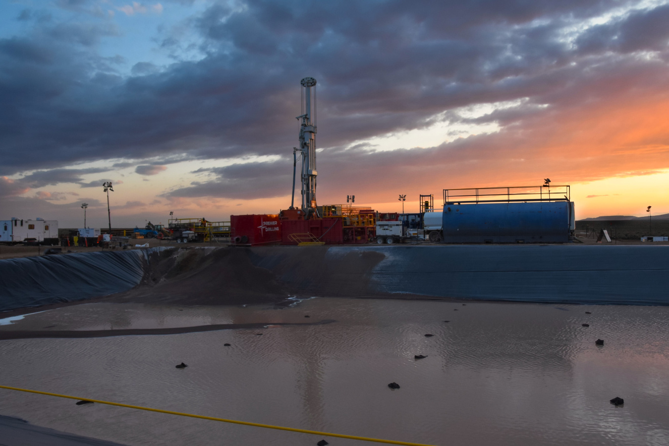 A large drilling well with machinery and trucks around it and a sunset in the background