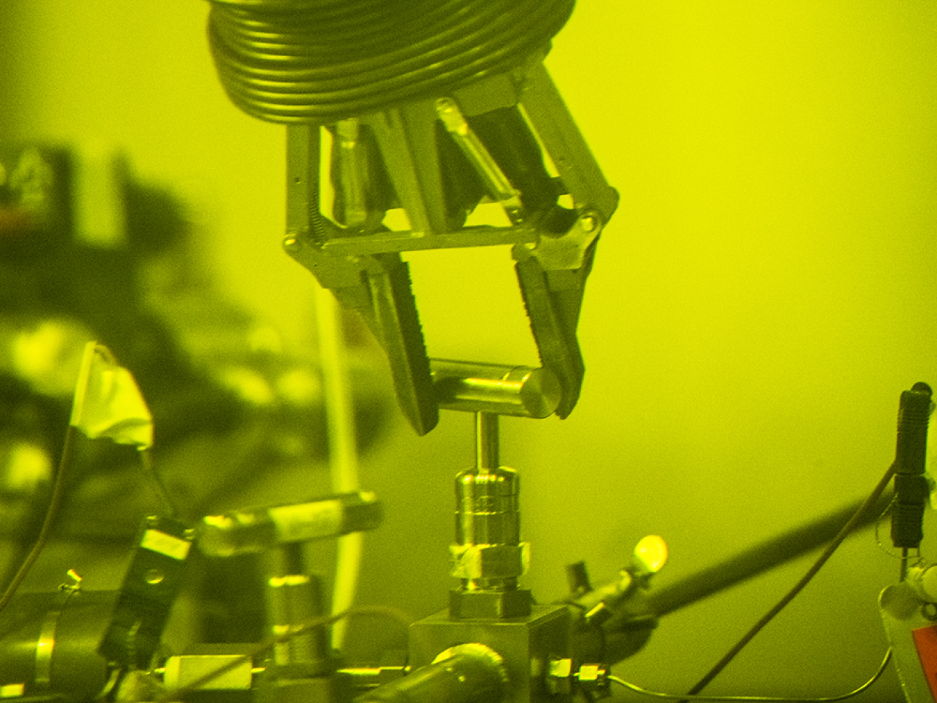 Green-tinted photo of a robotic claw holding a metal pin attached to lab equipment.