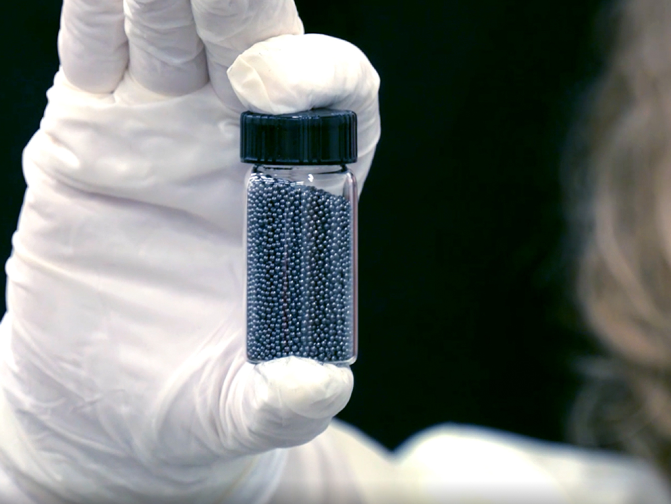 Gloved hand holding a bottle of small, round metallic TRISO pellets.