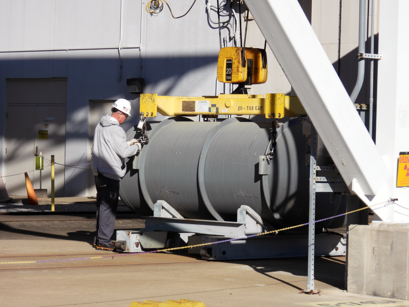 A large cylinder tank being lifted at the DUF6 center with a worker in safety gear standing beside it