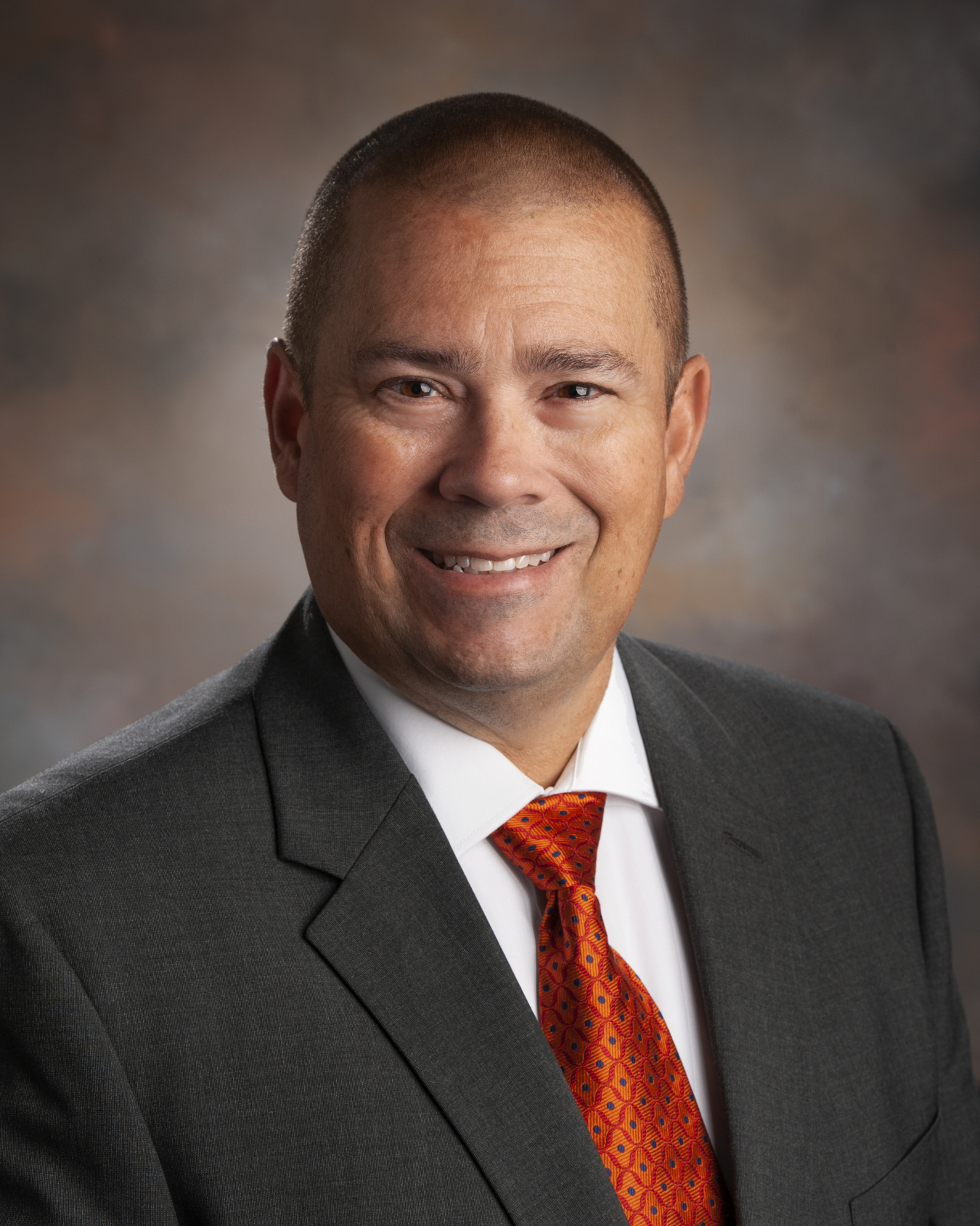 A headshot of a man in black suit with a red tie