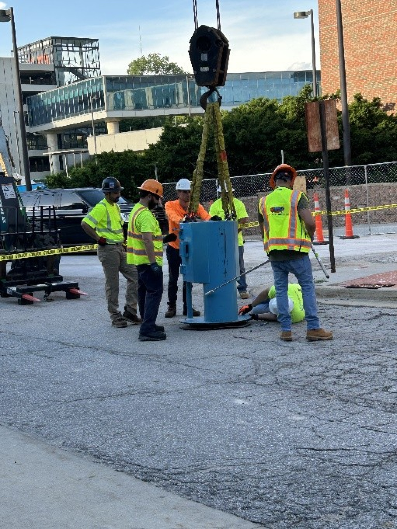 A group of people in high-visibility clothing stand in a parking lot around a blue cylinder with a large crane hook attached to it.