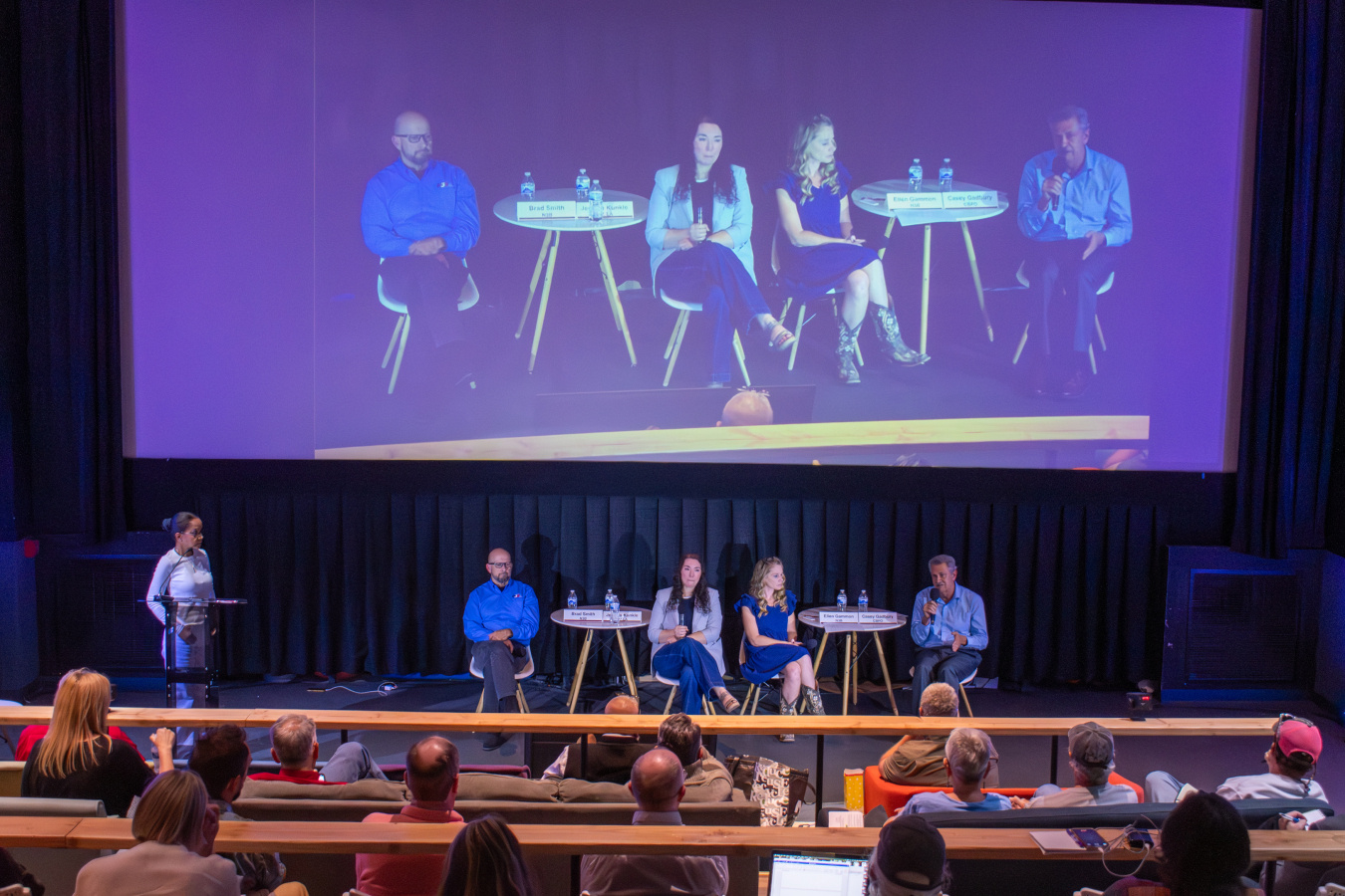 A group of people sitting on stage speaking to a crowd at a community forum