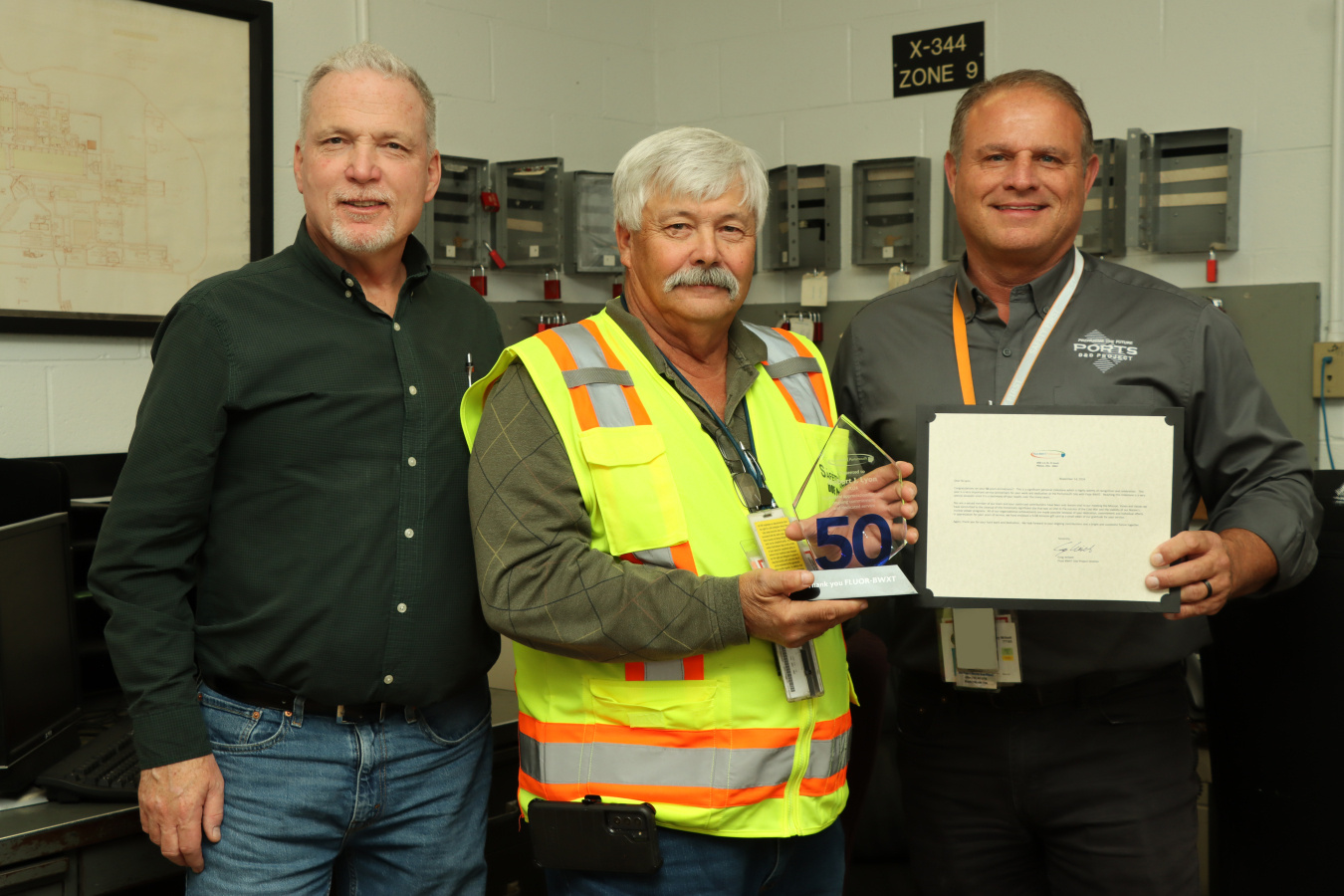 An employee at the Portsmouth Site in a yellow safety vest holding an award and posing for a group picture with two others