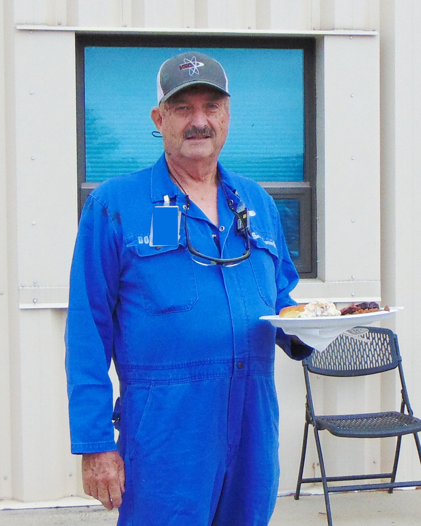 A smiling man in a blue work outfit and a grey hat holding a plate of food