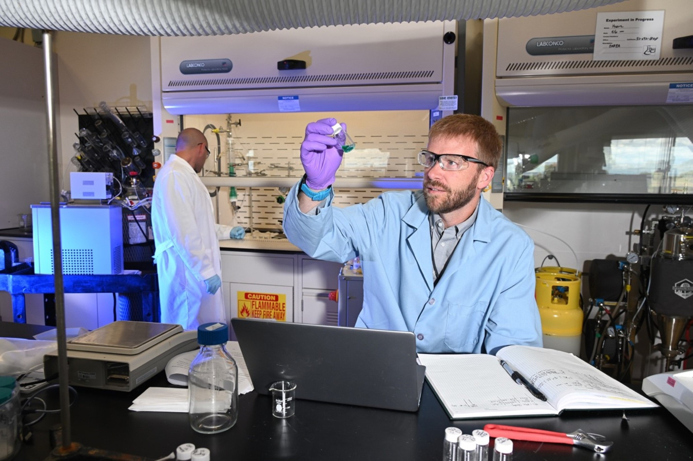 A man wearing a lab coat holds up a small vile filled with liquid in a laboratory room with lab equipment in the background. 