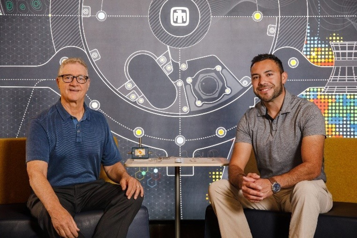 Two people seated in front of a wall of graphics next to a table.]
