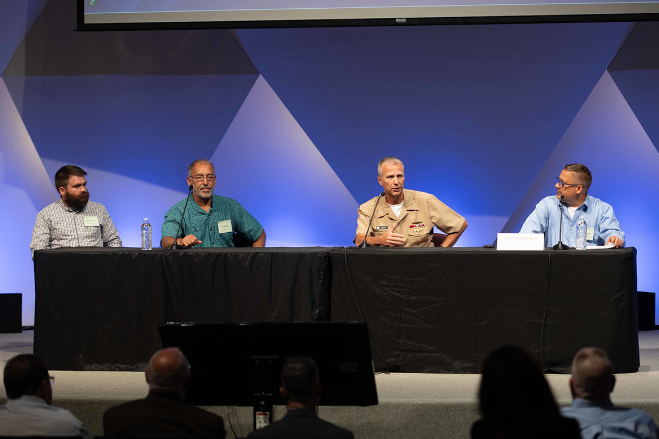 Four individuals sitting at a table on stage facing a crowd for a panel discussion session. 