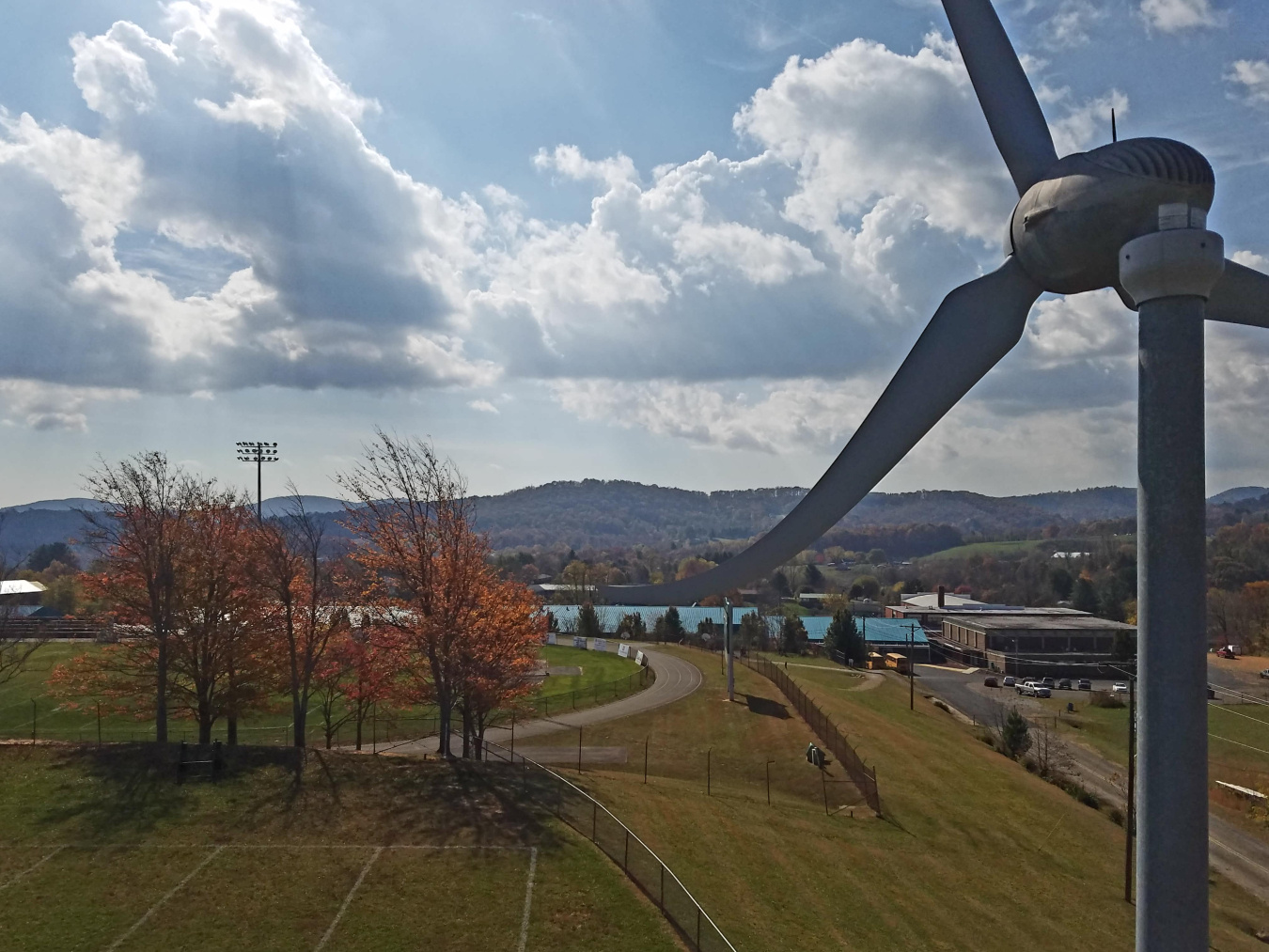 A small wind turbine overlooking athletic fields and a building in autumn mountains.