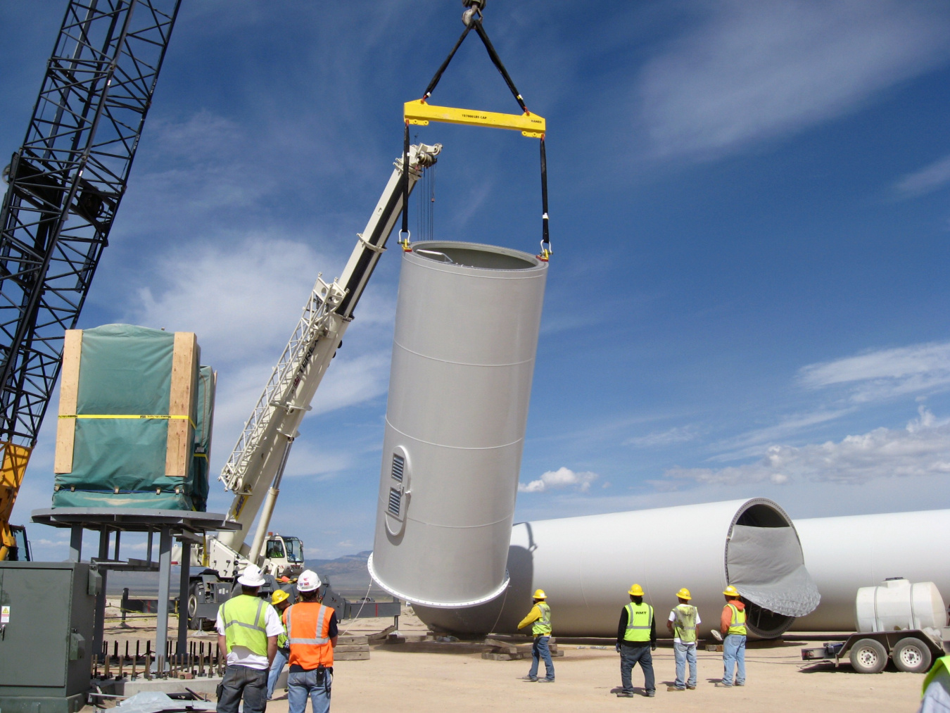 A crane lifting a large cylinder while workers in protective gear watch