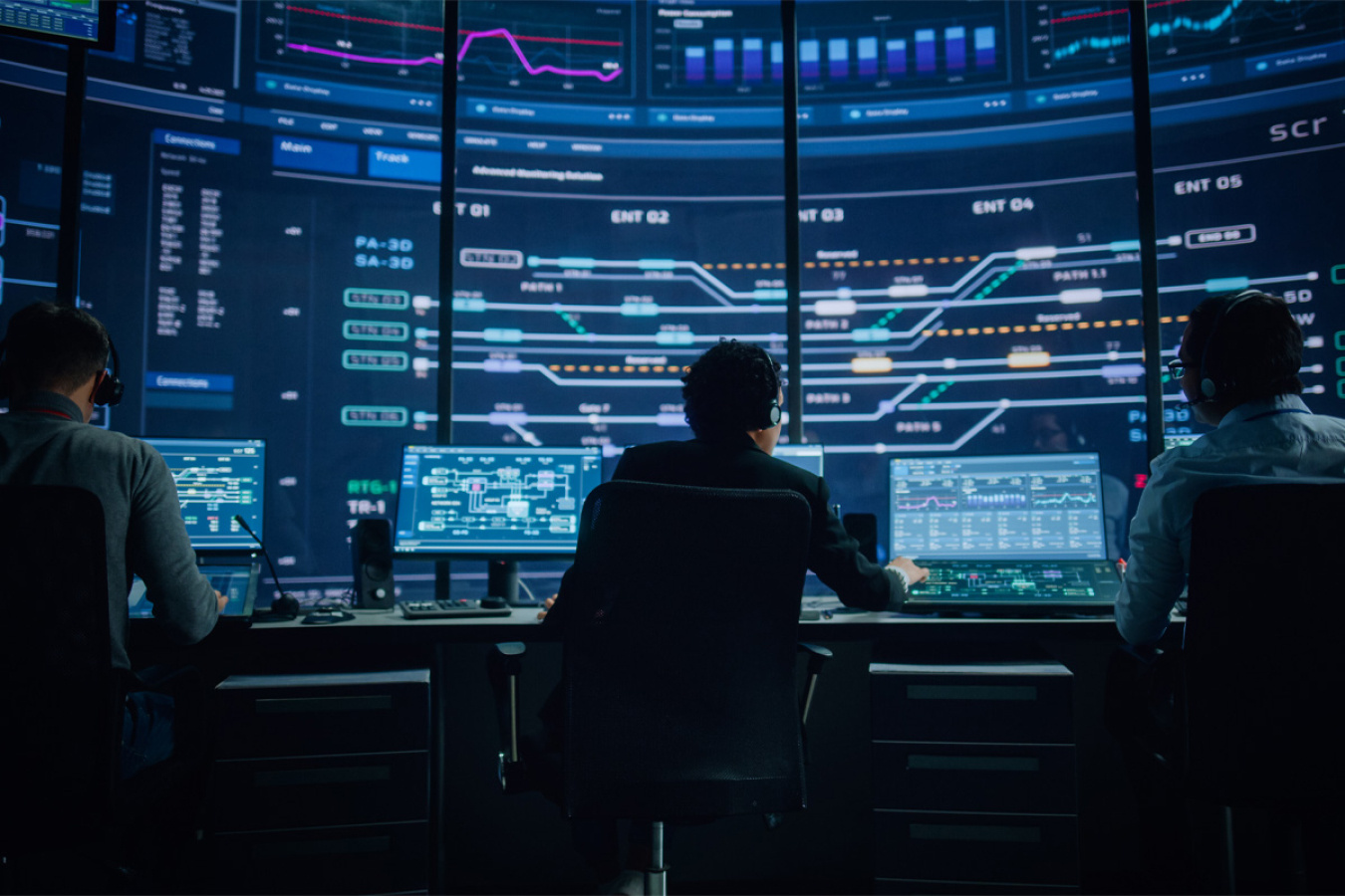 The back of a person is visible seated before three small monitors and huge monitors on the wall in front in a control operations center. 