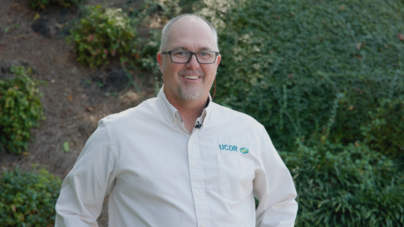 A professional headshot of a man with glasses in a tan button up shirt