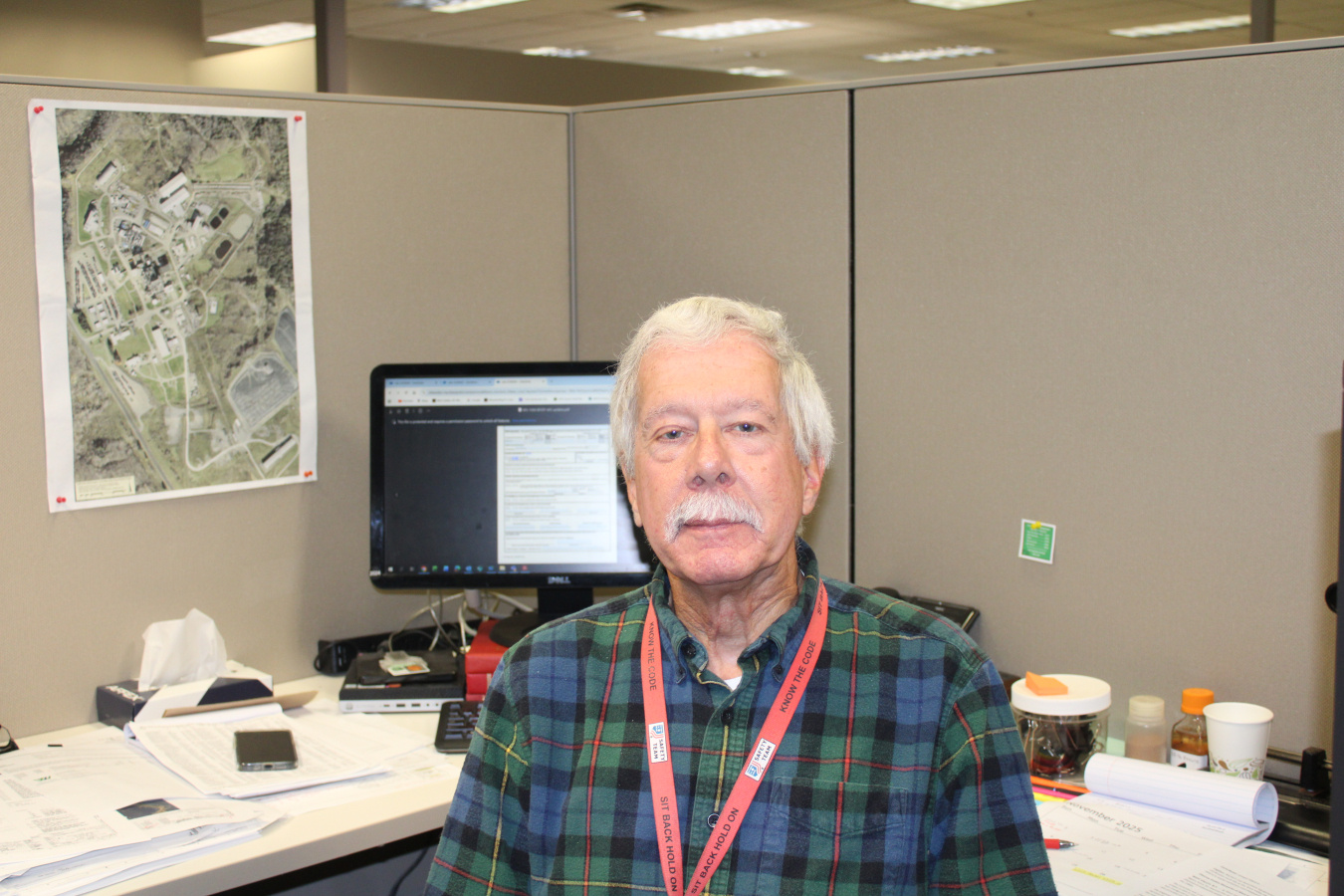 An older man in a blue and green plaid shirt sitting at his desk in a cubicle smiles for a photo