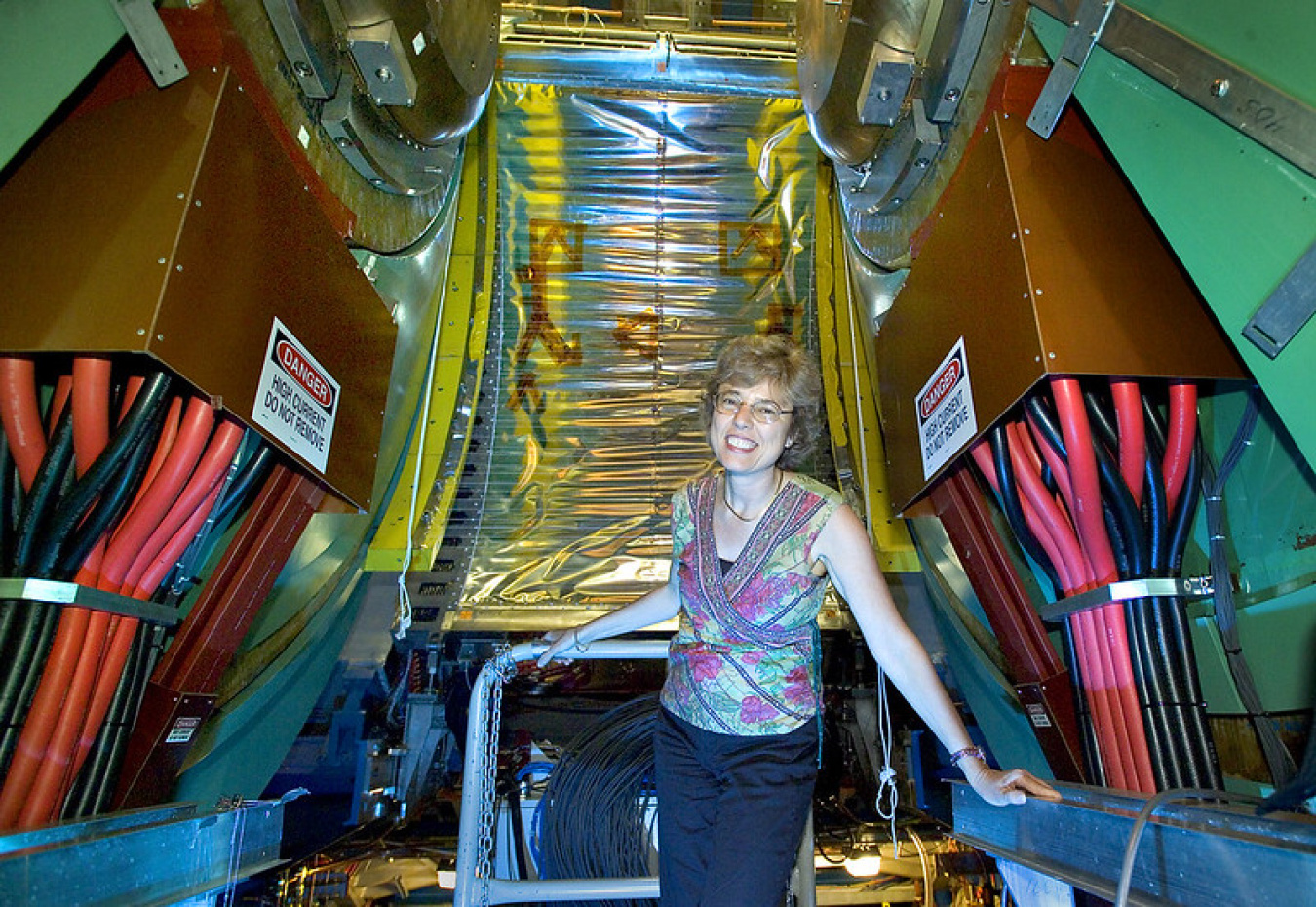 Barbara Jacak standing in front of the massive PHENIX detector. There are two huge copper-colored boxes with thick red and black wires running out of them labeled “Danger – High Current – Do Not Remove.” Directly behind her, the detector rises up with a silver/gold sheet of metal in the middle and yellow curving metal arms on either side. 