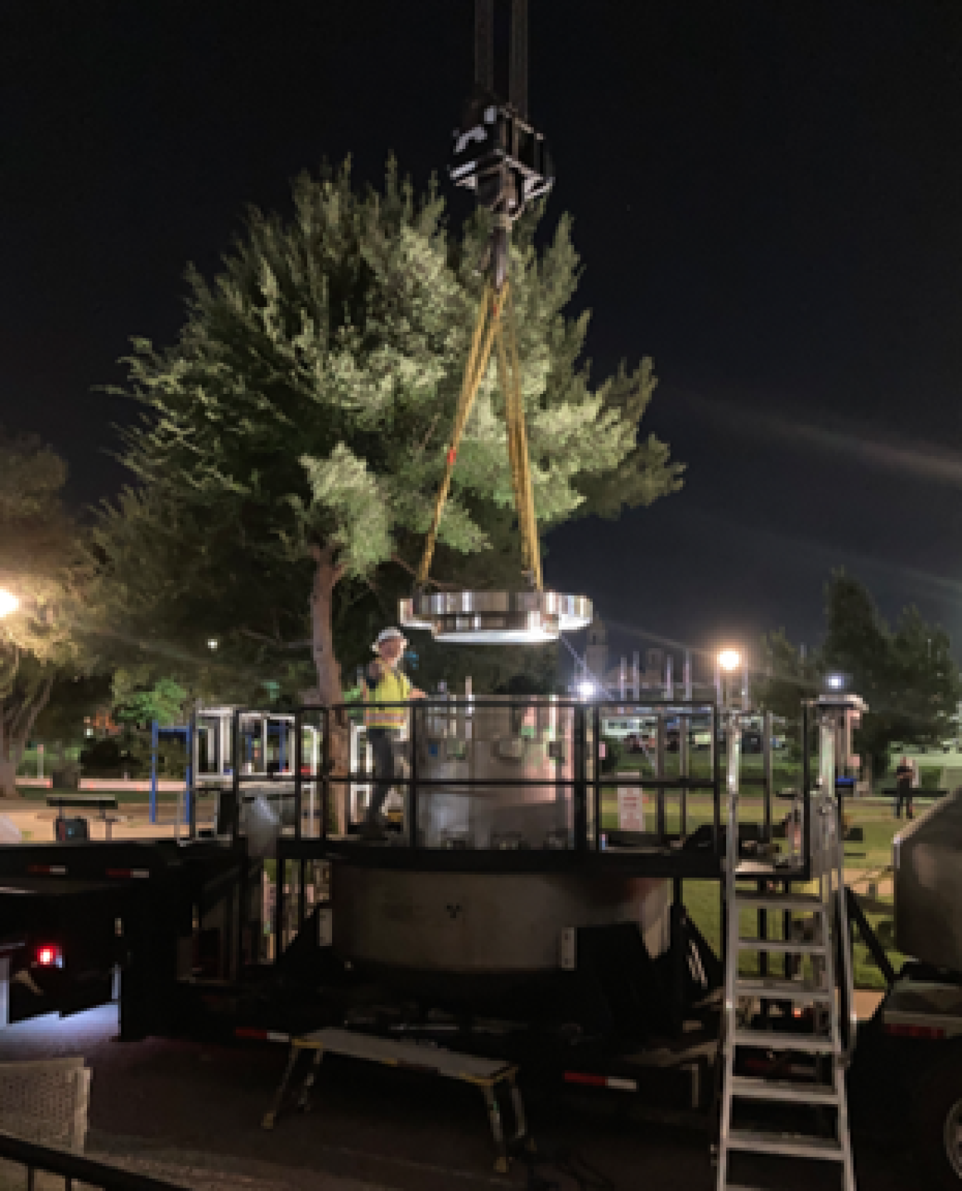 A man in a high-visibility vest guides a metal cylinder held by a crane onto a trailer in a parking lot. It's taking place at night and the scene is eerily lit by streetlights.