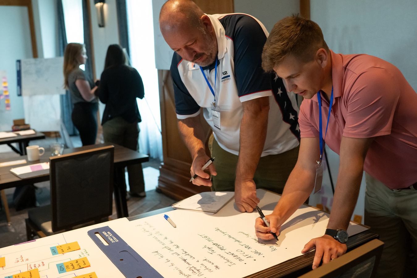 Two men use a large notepad to document ideas and information with other teams collaborating in the background. 