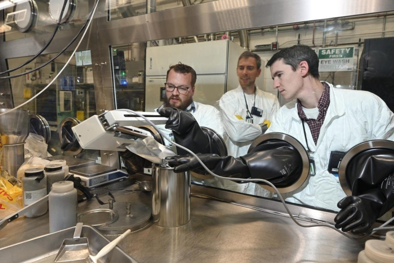 Three scientists wearing lab coats operate a glove box