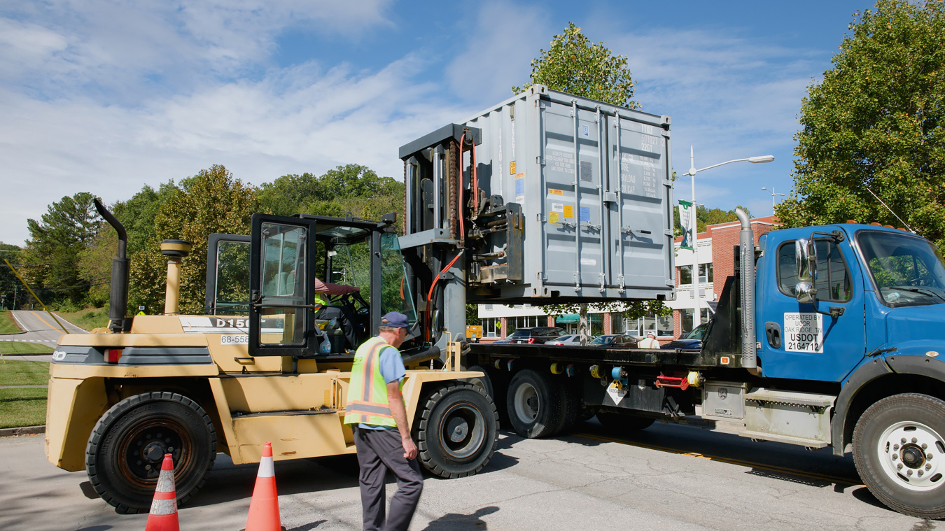 A truck hauling a hot cell and a construction vehicle beside it with an employee in safety gear walking along it
