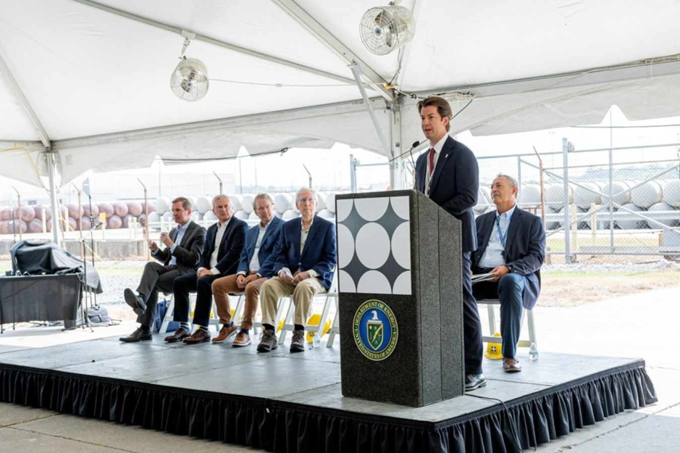 A man standing behind a podium giving a speech with five men sitting in chairs on the stage behind him