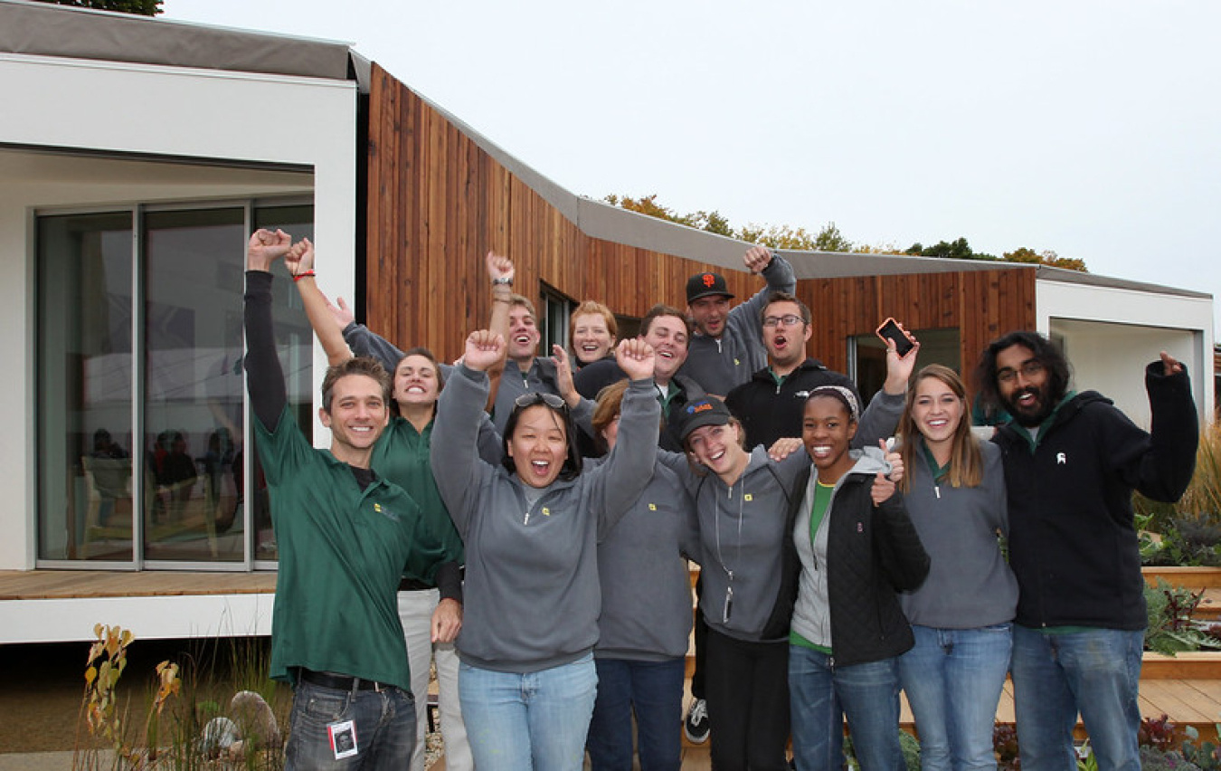 A group of students standing in front of their competition house, facing the camera and joyfully raising their fists in the air.