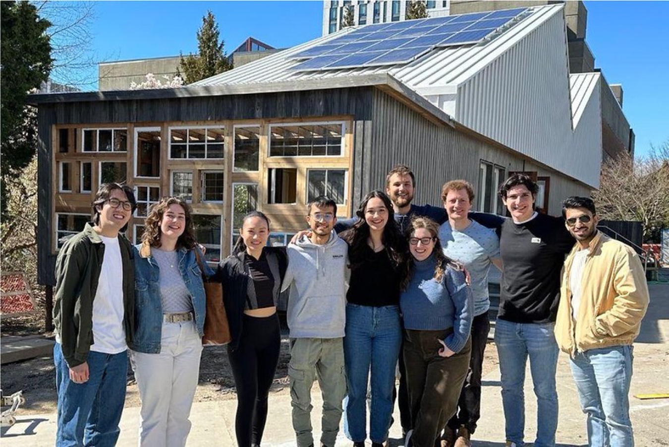 10 people standing in a row facing the camera (this university team) with the team's house in the background.