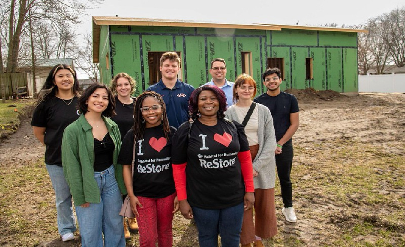 Nine people (this team) standing together facing the camera, with their house under construction behind them.