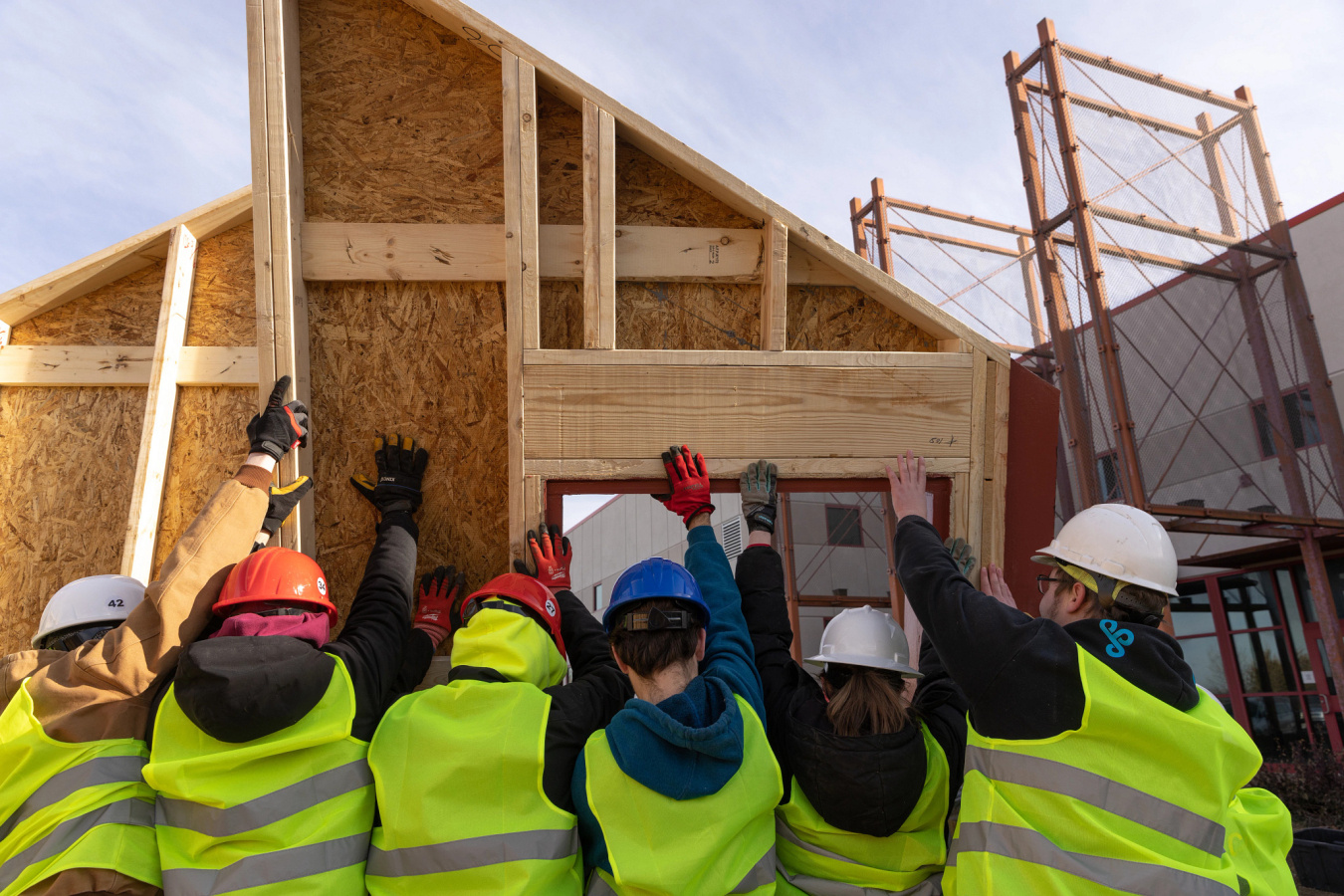 A crew of student workers in hard hats and construction vests, their backs to the camera, lifting a house structural panel in place.