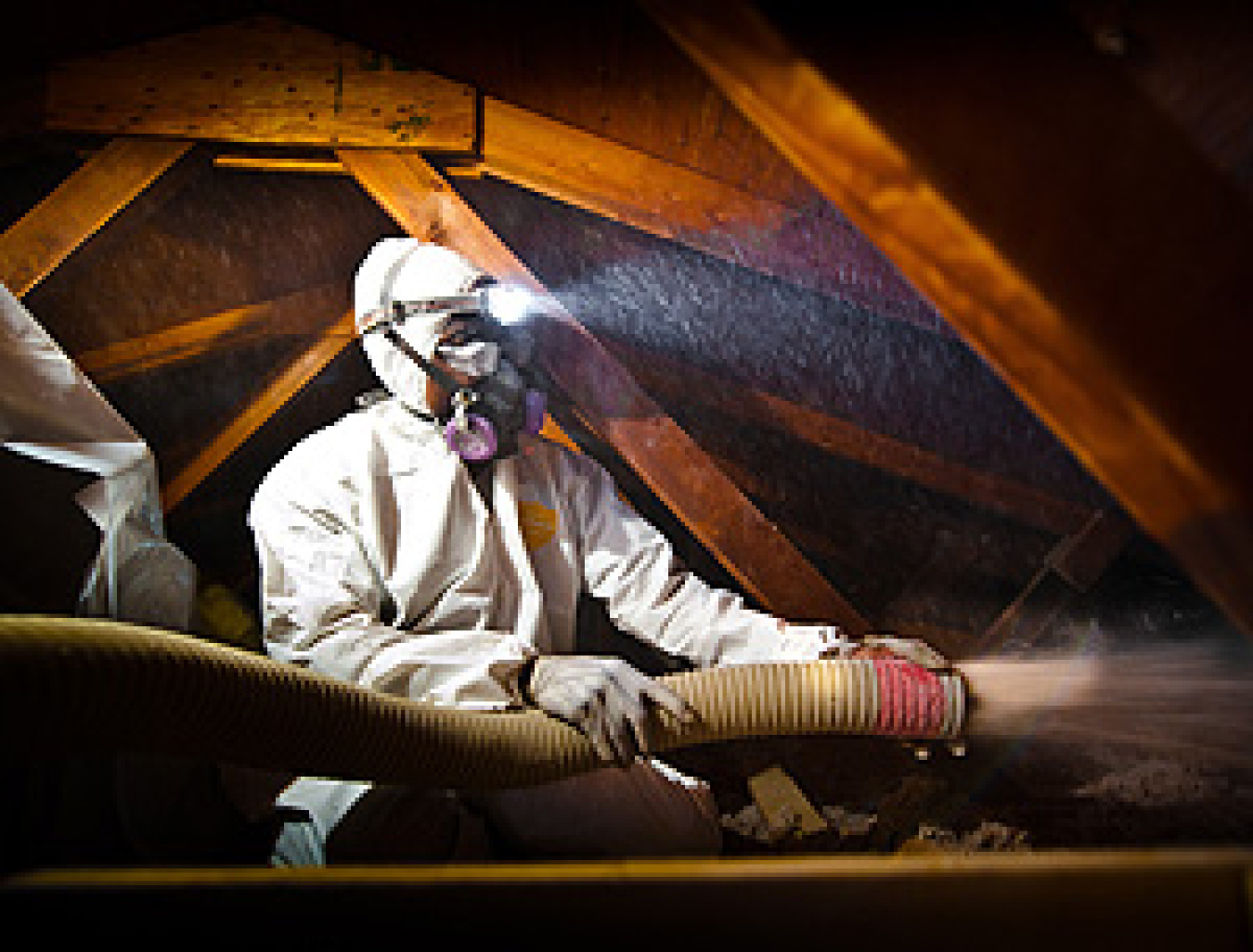 Worker in a protective suit in an attic, with rafters above, using equipment to blow in insulation.