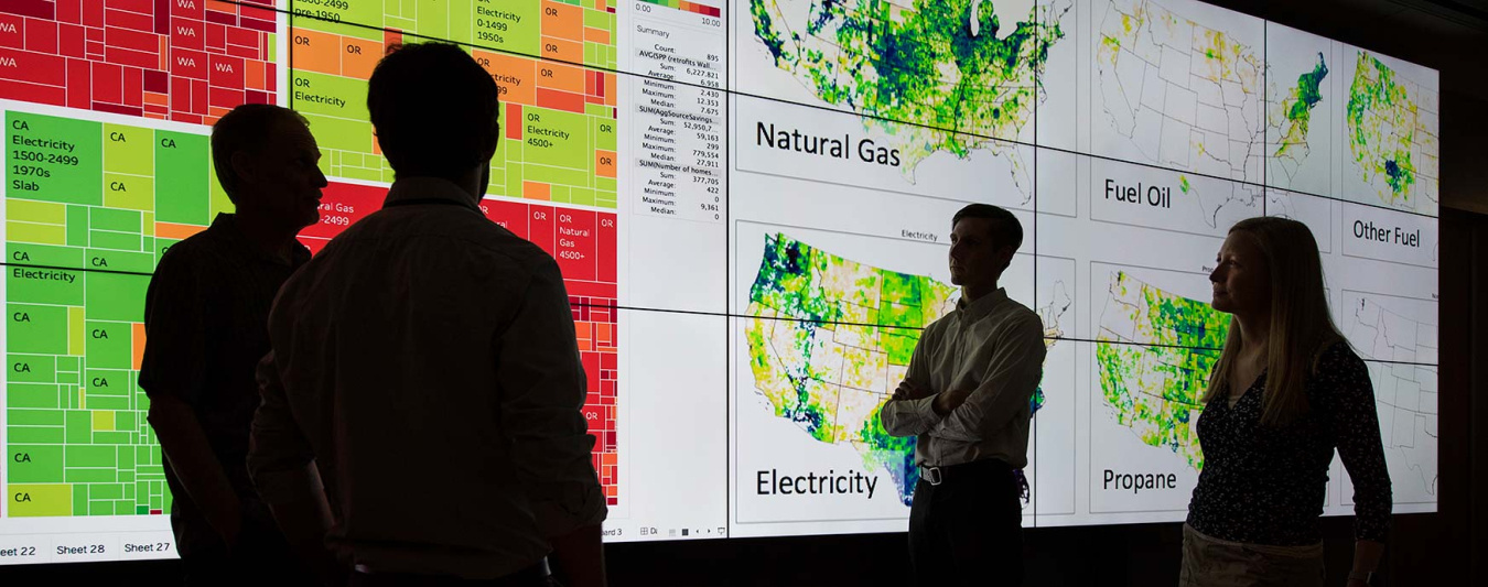 Four people silhouetted in front of a large display screen with maps and other data.