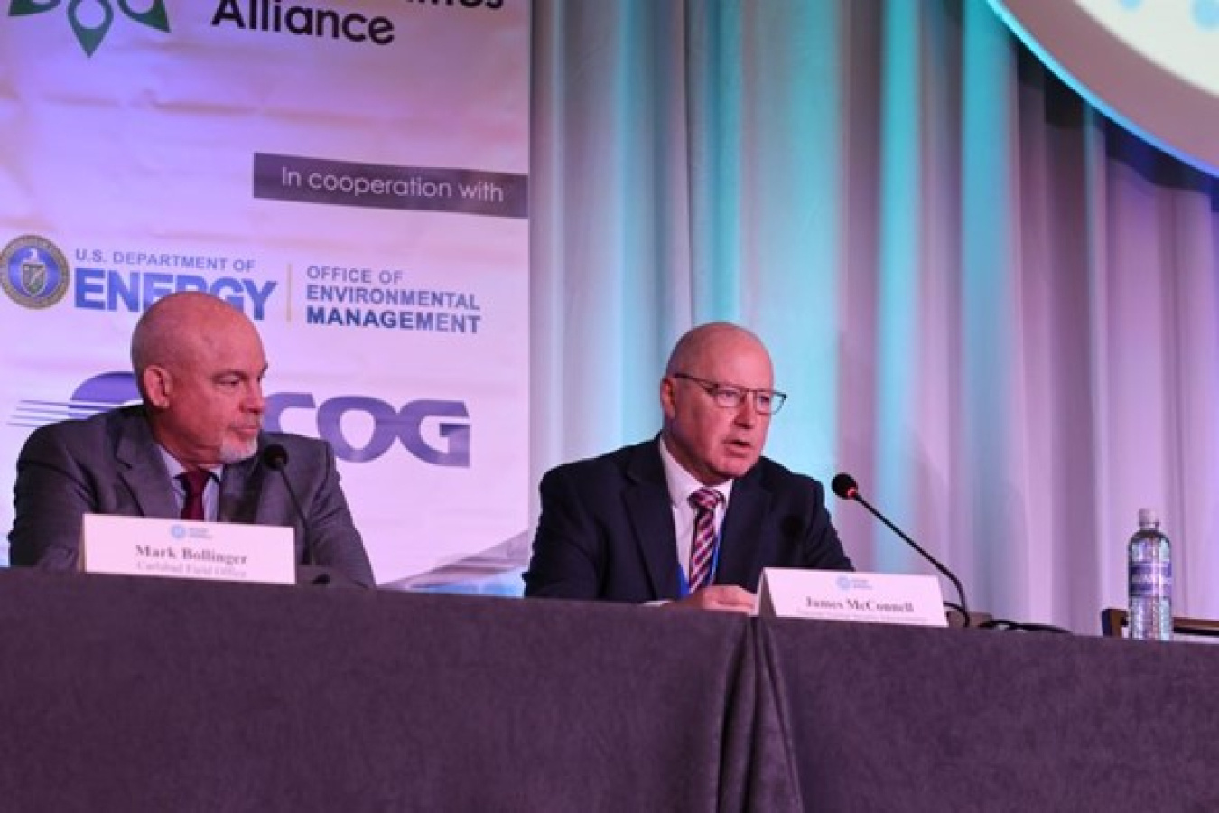 Two men sit behind a covered table. One man is speaking into a microphone.