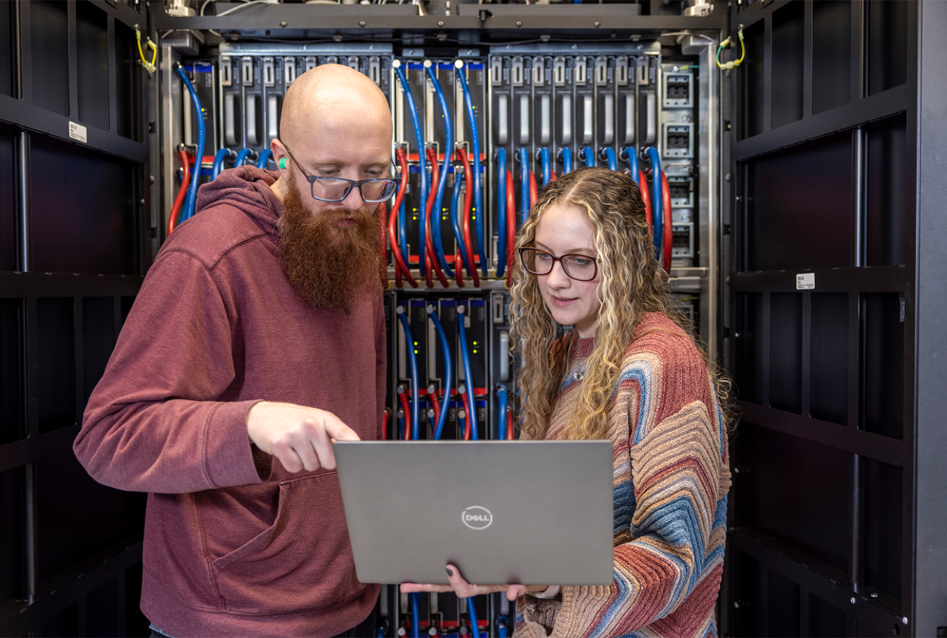 Two researchers stand facing a laptop computer with a server rack in the background.