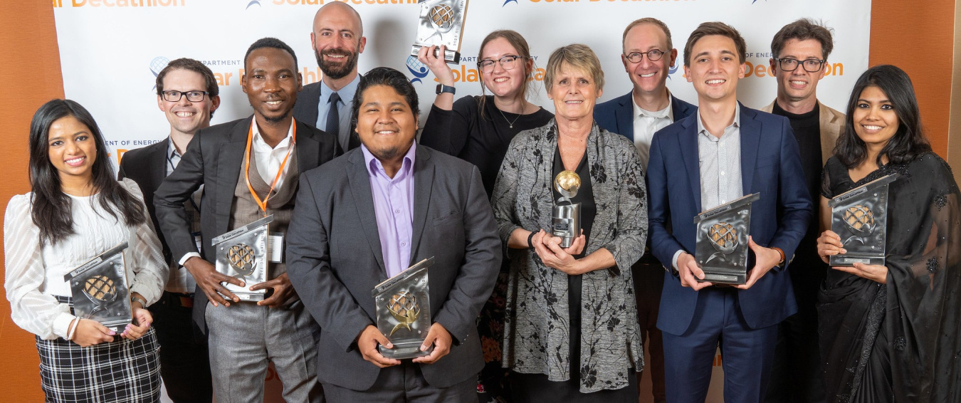 Group of 11 people in business attire standing together, facing the camera, and many of them can be seen holding awards.