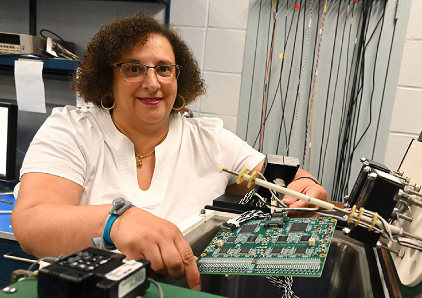 Mary Bishai (a woman in a white t-shirt) sitting at a desk with a microelectronics board in front of her and electronics equipment to the side.
