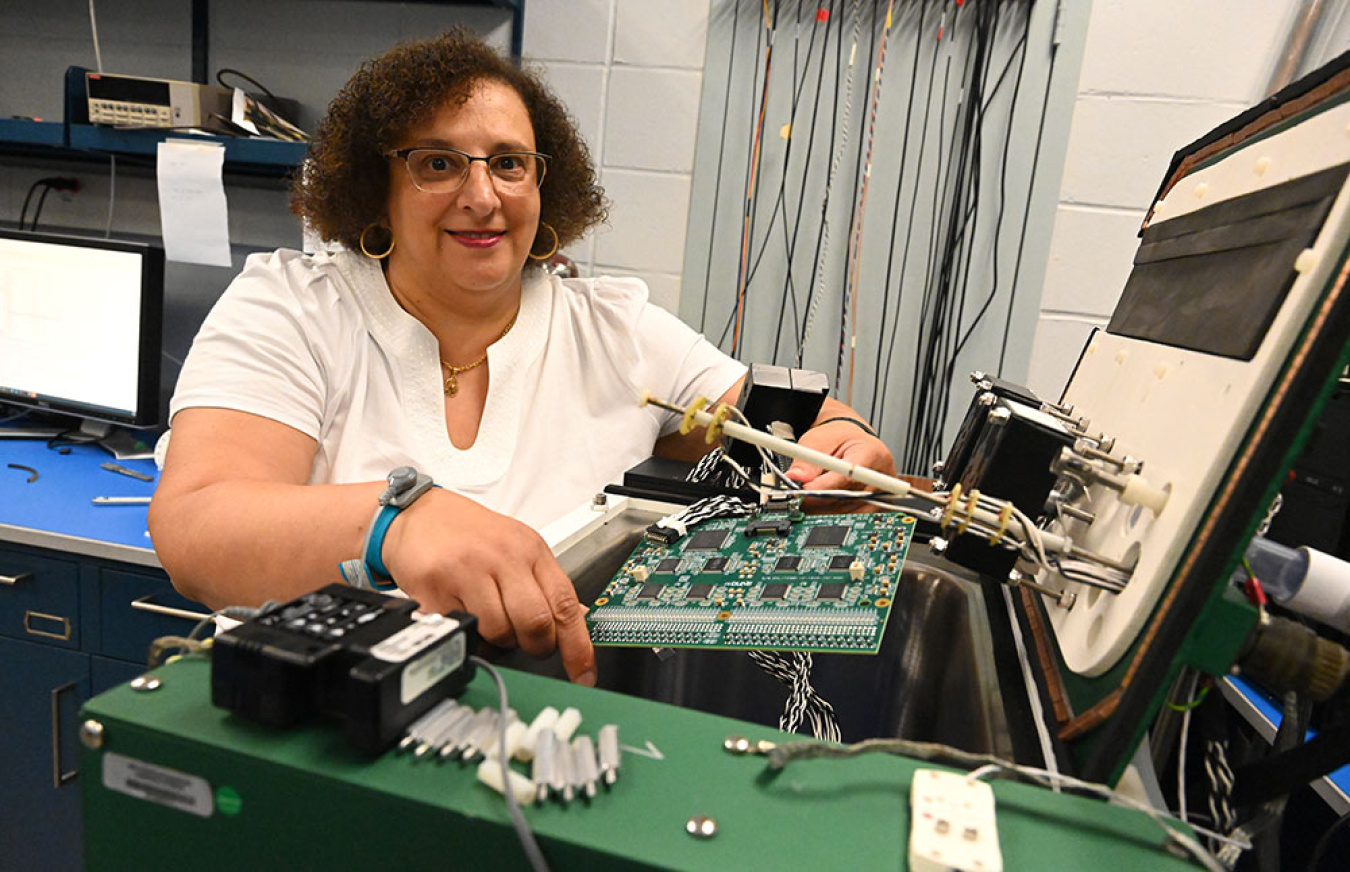 Mary Bishai (a woman in a white t-shirt) sitting at a desk with a microelectronics board in front of her and electronics equipment to the side.