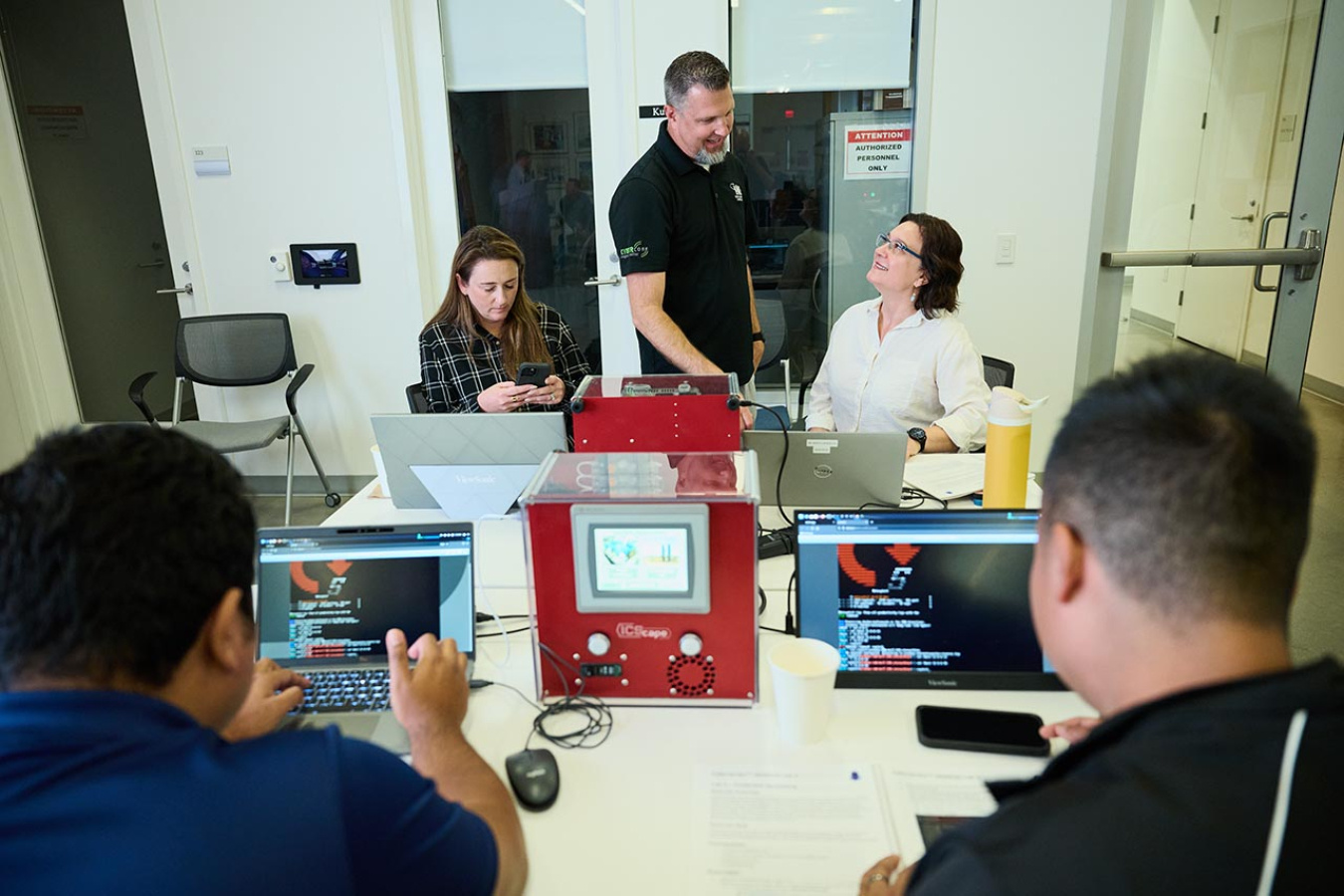 Four people seated at desks conducting cyber training on laptops. 