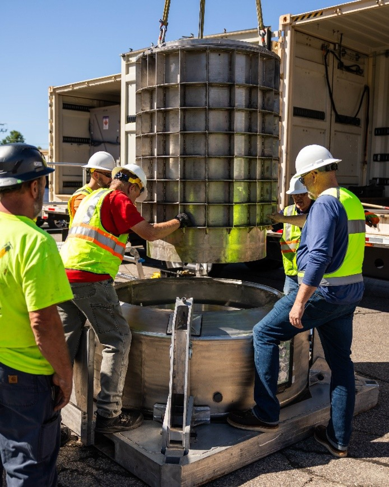 People in high-visibility clothing and white hardhats help lower a cylinder into a large base.