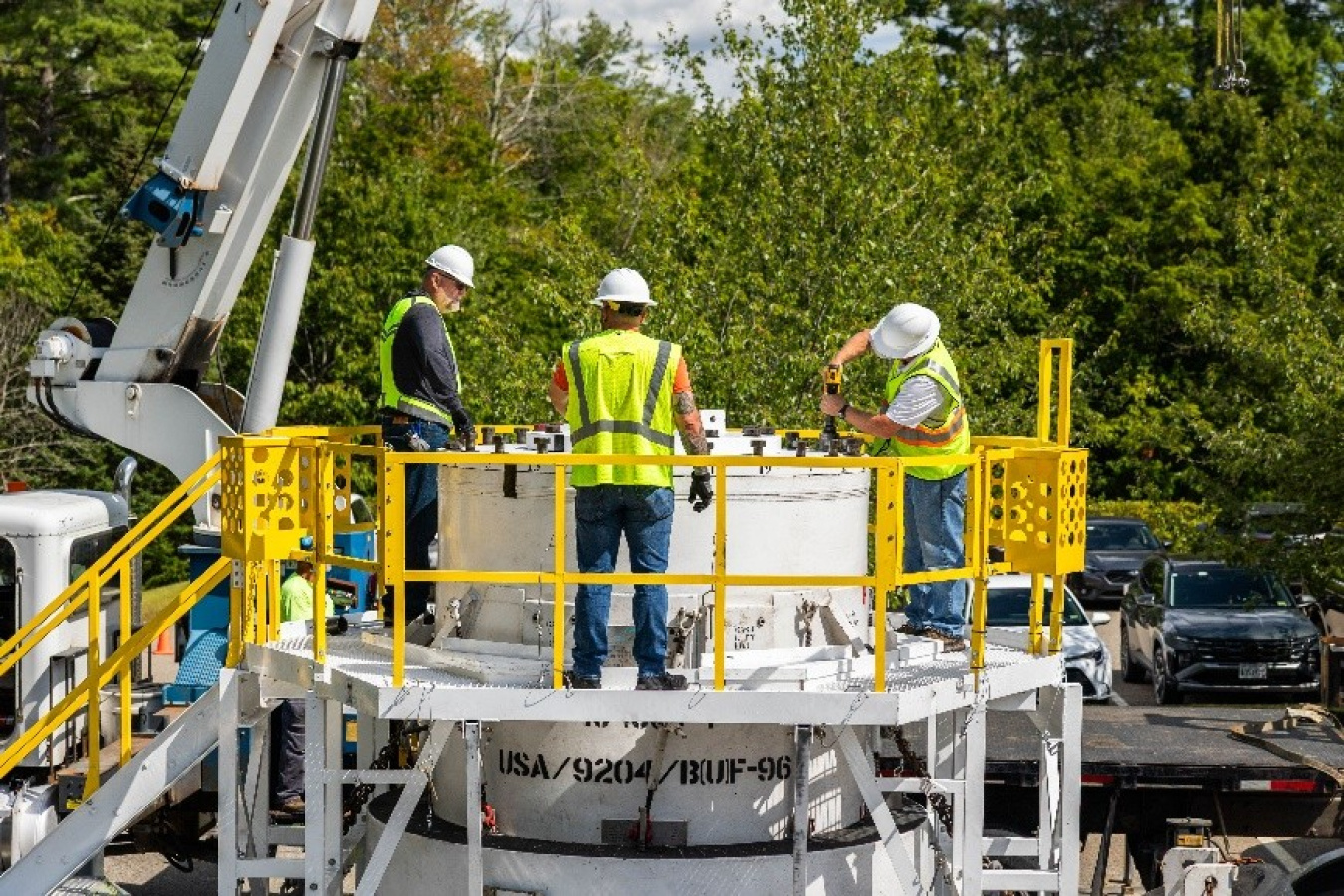 Three people wearing high-visibility clothing and wearing white hardhats stand on a platform and prepare a cylindrical base that will hold a removed irradiator for transport.