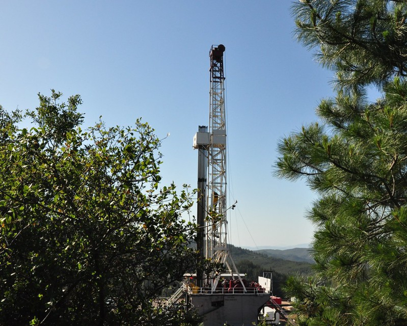Photo of a drill rig between trees in the foreground, with rolling hills extending into the background.
