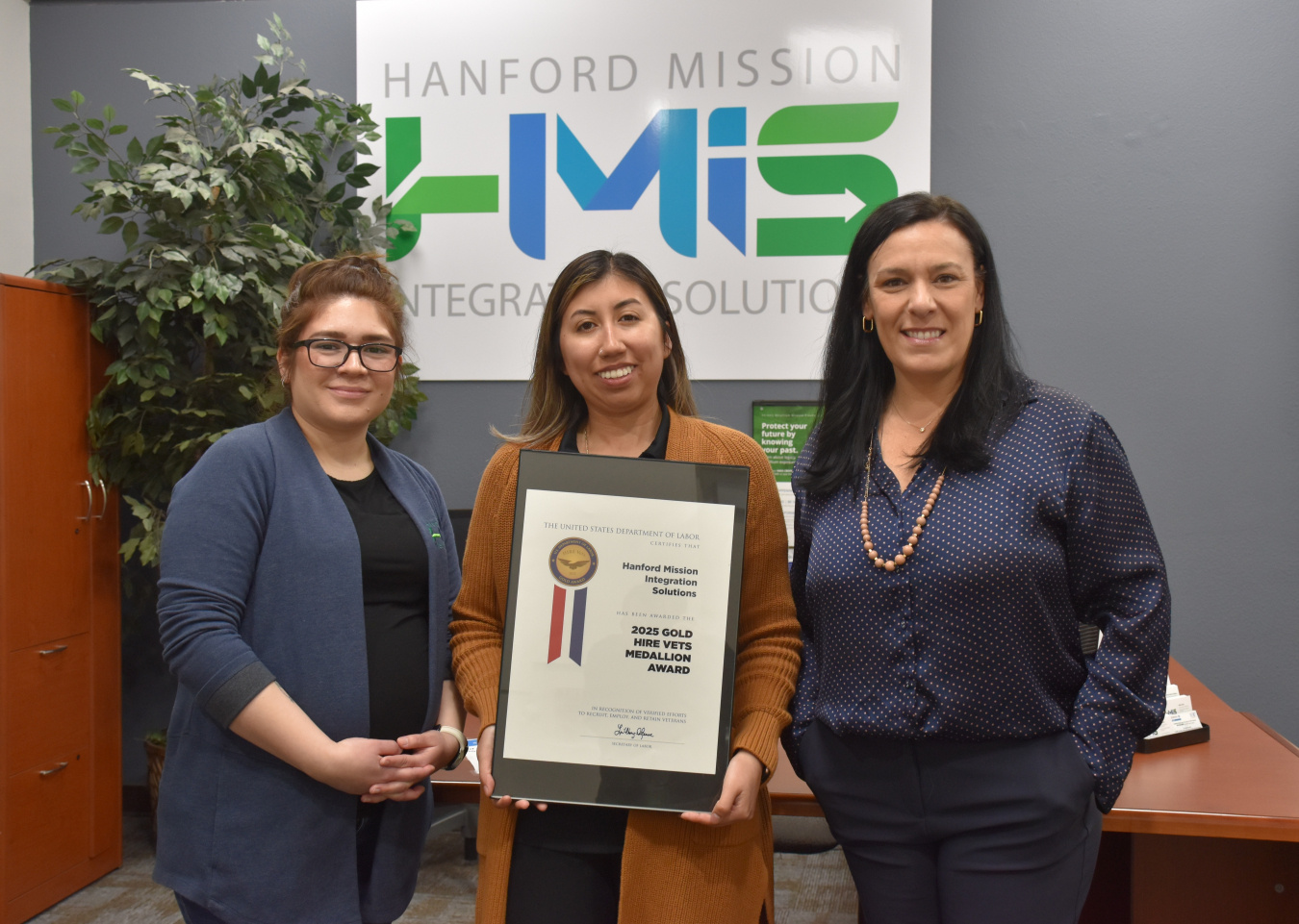 Three women posing for a group picture, the one in the middle is holding a certificate 