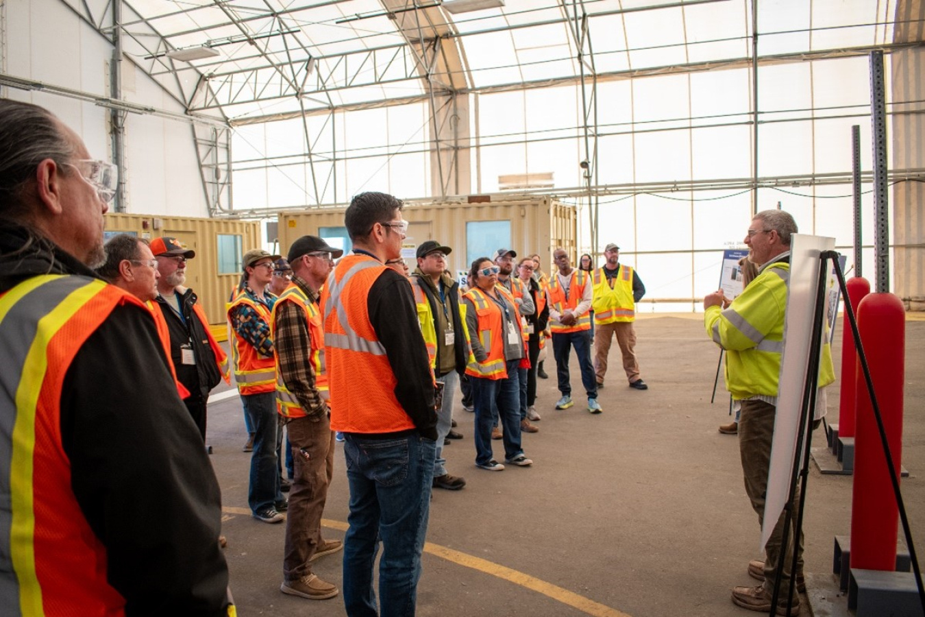 A group of people in yellow and orange safety vests listening to a man talk at the front of a room