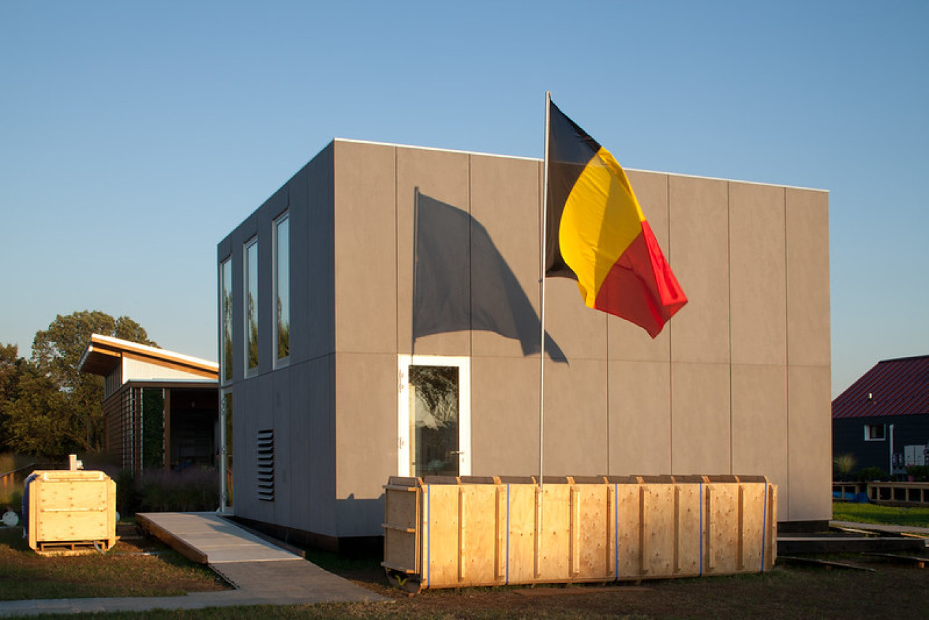 Exterior of Team Belgium's Build Challenge house, a simple square structure with a door on the side facing the camera, windows on the other visible side, and a flag flying  next to it.