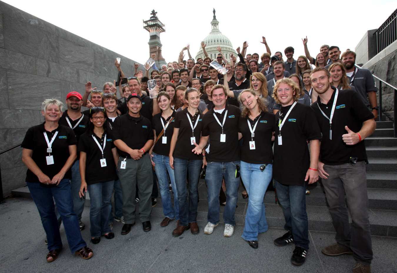 A group of students wearing matching shirts and lanyards standing together, facing the camera, in an outdoor setting with the dome of a Capitol / government building in the distance behind them.