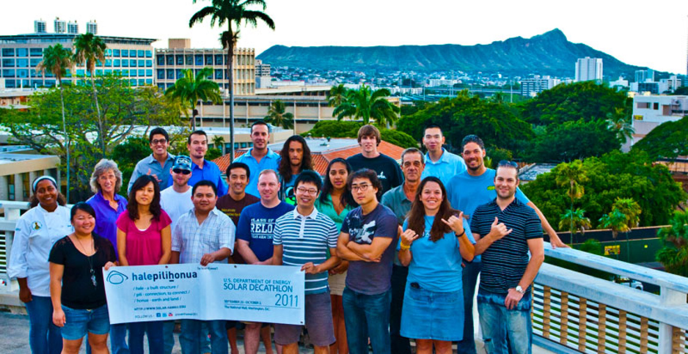 Group photo of the University of Hawaii team, with a few members standing in the front of the group holding a sign, and there are some buildings and mountains of Hawaii in the background.