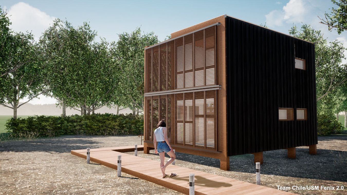 Exterior of the Chile team's Build Challenge house, a two-story structure with grate windows on the front and a board sidewalk in front of it, with a woman standing on the sidewalk in front of the house.