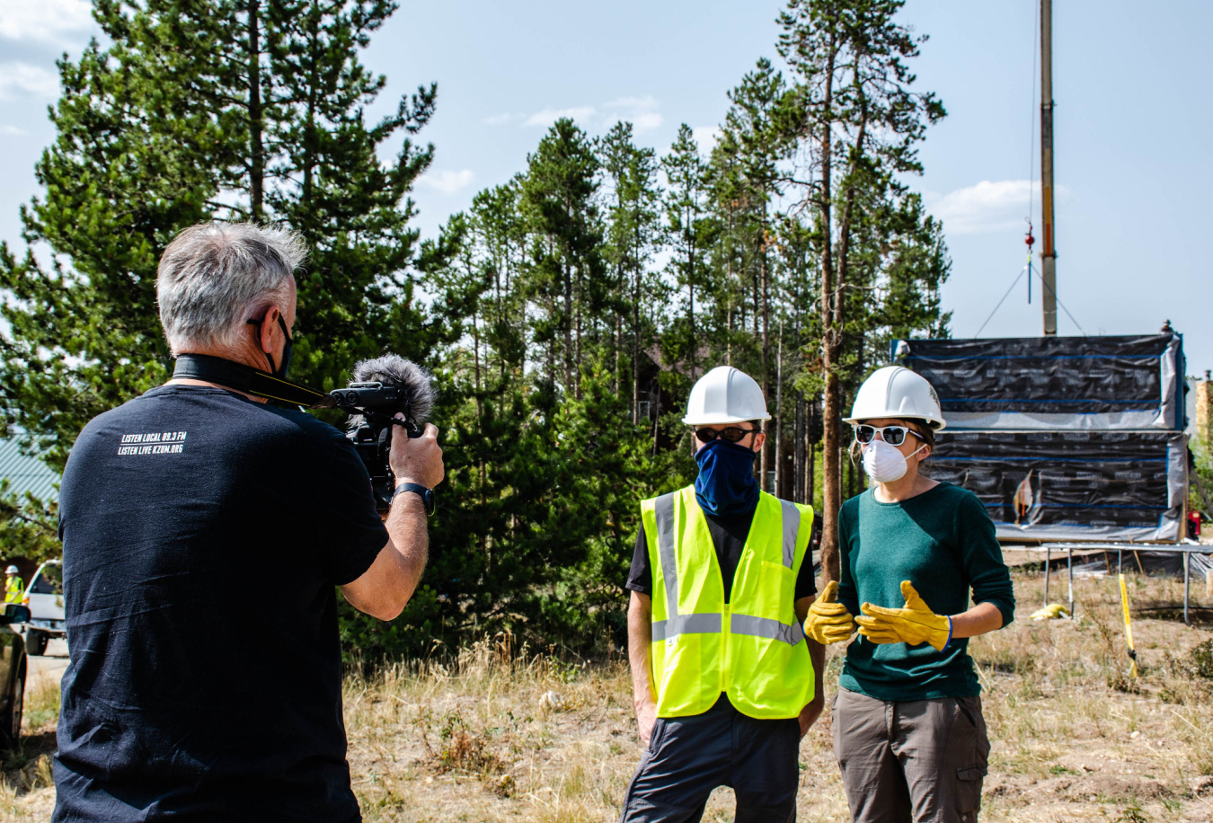Three people standing near a Build Challenge competition house site, two of them in hard hats, facing the other person, who is taking their picture.