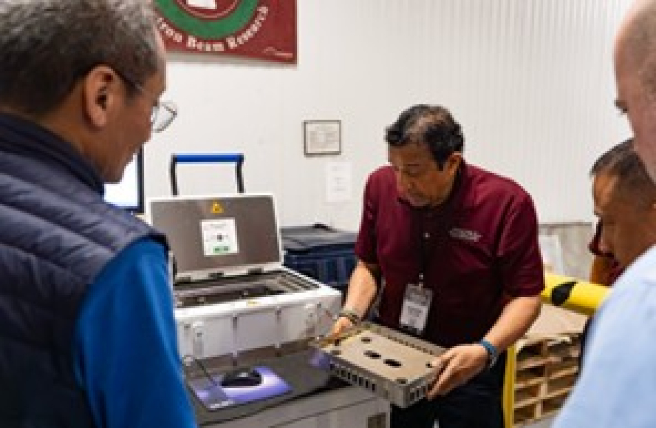Standing next to a machine, a man holds something looking like a tray. Three people look on.